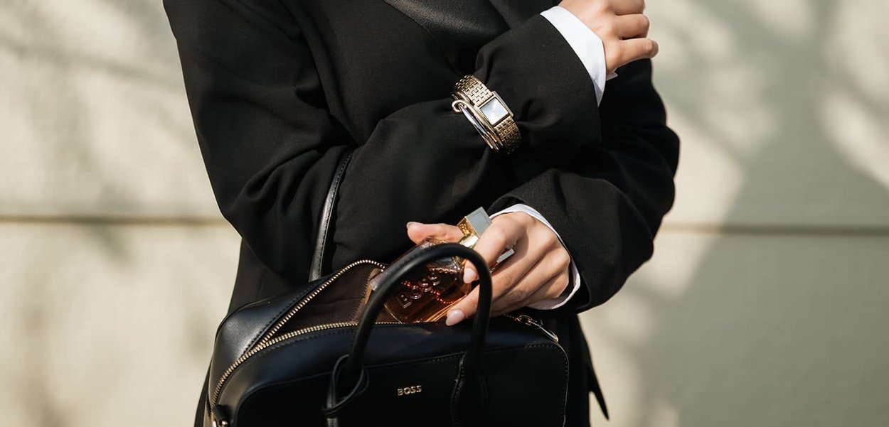Persona con blazer negro sosteniendo un frasco de perfume y llevando un bolso negro BOSS con cremallera dorada y reloj.