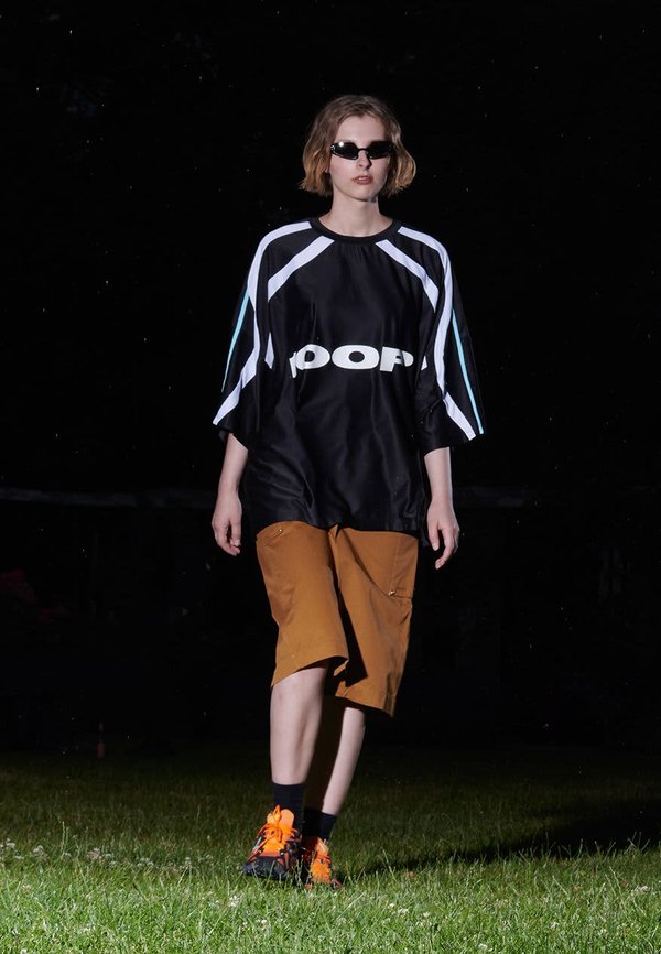 Camisa negra de gran tamaño con detalles en blanco y azul, combinada con pantalones cortos marrones y zapatillas naranja vibrante. El modelo lleva gafas de sol negras.