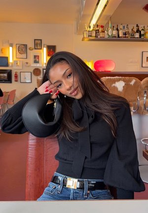 Schwarze Handtasche mit strukturierter Oberfläche und geschwungenem Design, entspannt gehalten. Modell trägt ein schwarzes Oberteil mit Schleifendetail und blaue Jeans.