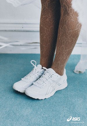 Jambes portant des collants marron à motifs et des chaussures de course blanches Asics, se tenant sur un tapis bleu clair près d'un mur blanc.