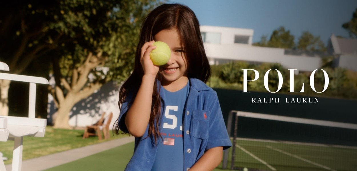 Joven niña sonriendo en la cancha de tenis sosteniendo una pelota de tenis amarilla cerca de su rostro, vistiendo una camisa azul Polo Ralph Lauren.