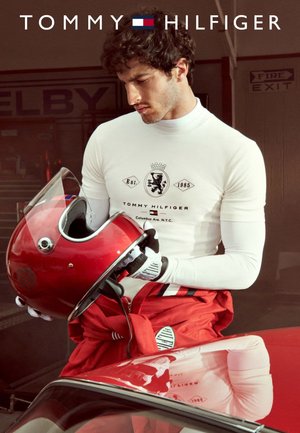 White turtleneck with black logo, red racing gear tied at waist, and a red helmet with visor. Vehicle in foreground, garage background.