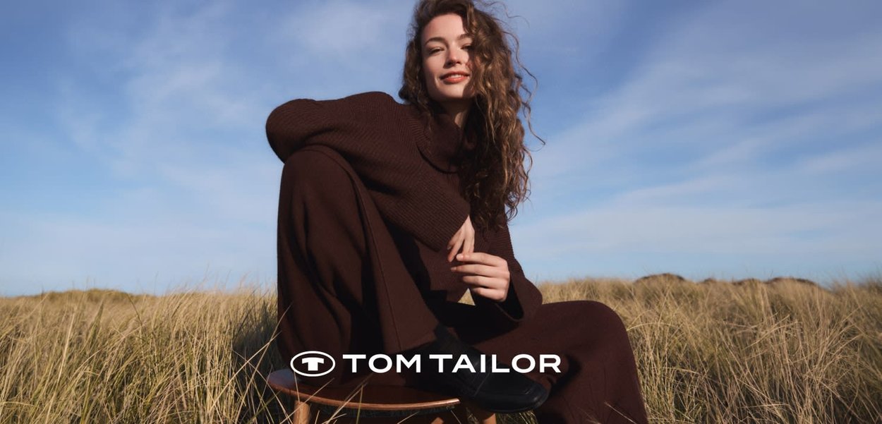 Młoda kobieta w brązowym swetrze siedząca na drewnianym stołku na polu trawy pod niebieskim niebem, delikatnie się uśmiechając.