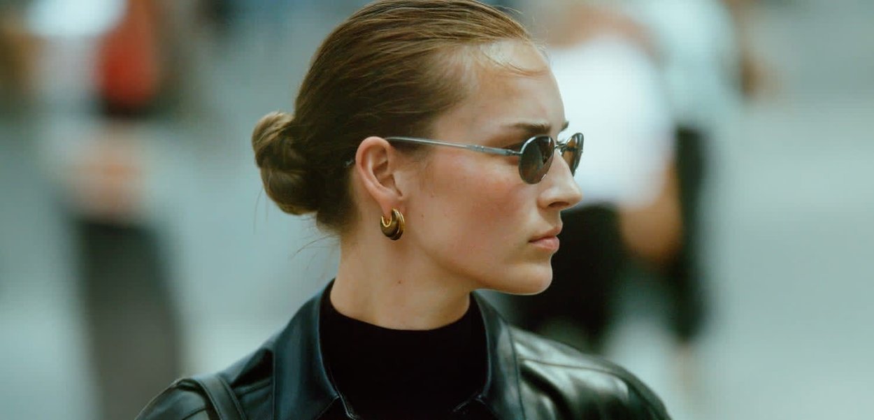 Veste en cuir noir, haut à col montant, boucles d'oreilles en or, et lunettes de soleil sombres. Cheveux lisses coiffés en chignon bas, vue de profil.