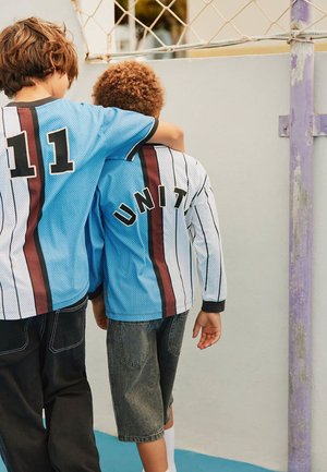 Twee jongens in sportshirts staan naast elkaar, waarbij de ene zijn arm om de schouder van de andere legt, vlak bij een basketbalring.