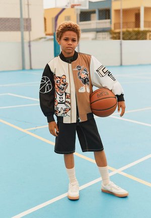 Junge hält Basketball auf einem Außenplatz, trägt eine bunte Jacke mit Tiger- und Vogelmotiven, schwarze Shorts, weiße Socken und Turnschuhe.