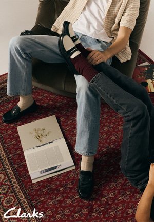 Chaussures noires avec accents blancs, semelle épaisse ; jean en denim ; chaussettes bordeaux ; tapis texturé ; magazine au sol ; cadre intérieur détendu.