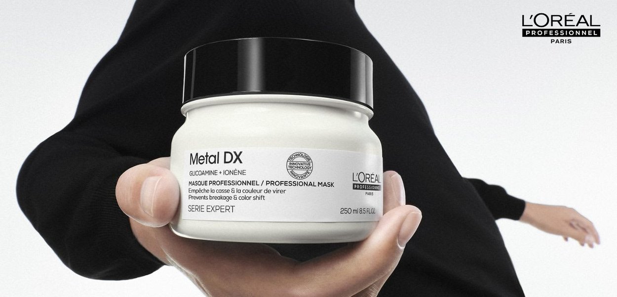 Ręka trzyma słoik profesjonalnej maski L'Oréal Metal DX z czarną pokrywką na białym tle, z logo L'Oréal w prawym górnym rogu.