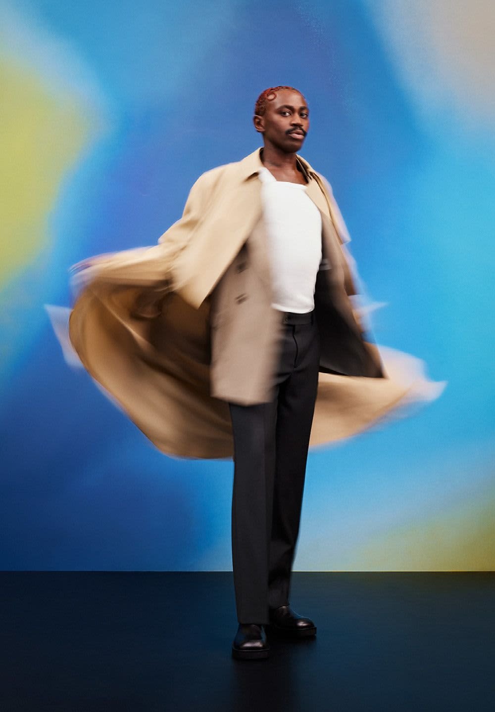 Beige trench coat med flytande design, bärs över en vit skjorta och mörka byxor. Svarta skor. Livlig blå och gul bakgrund.