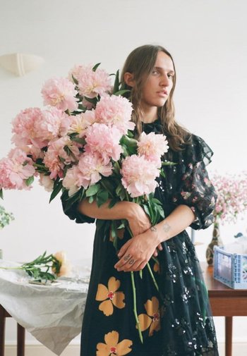 Sort gennemsigtig kjole med blomsterdetaljer, holder en stor buket af lyserøde pæoner med grønne blade; tatoveringer synlige på armene.