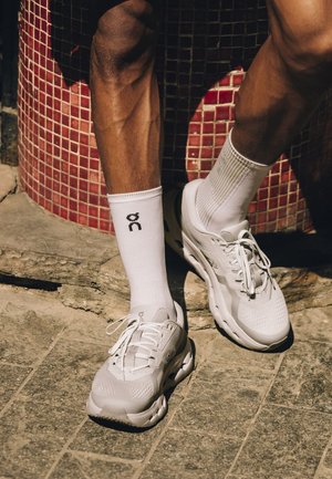 Gambe con calzini bianchi a coste con logo e scarpe da ginnastica bianche che si trovano su un pavimento in pietra vicino a una struttura con piastrelle di mosaico rosse.