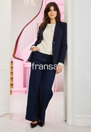 Frau lehnt an weißer Wand, trägt dunkelblazer, cremefarbene Bluse, dunkelblaue Marlene-Jeans und spitze braune Schuhe, im Hintergrund eine rosa Wendeltreppe.