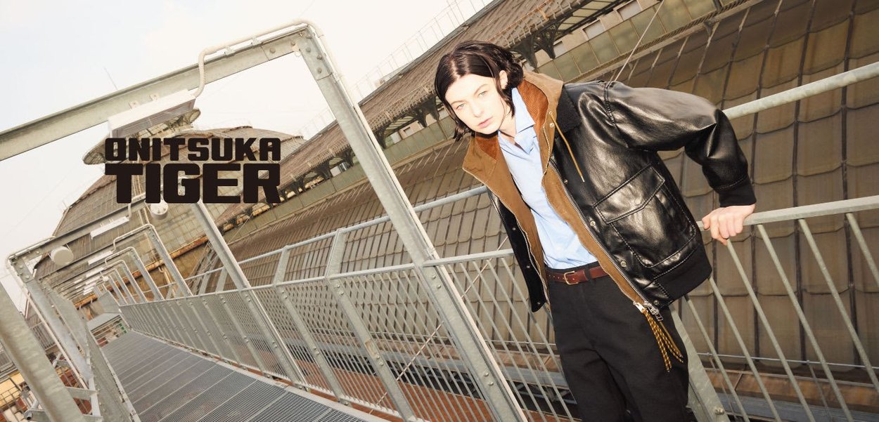 Czarna skórzana kurtka z brązowym kołnierzem, na niebieskiej koszuli i czarnych spodniach. Metalowa barierka i przemysłowe tło w stonowanych kolorach.