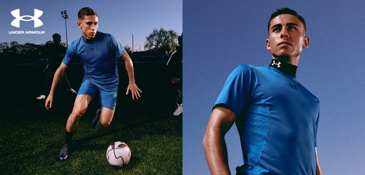 Blaues Sportshirt und Shorts aus feuchtigkeitsableitendem Material, mit hohem Kragen und Under Armour-Logo. Ein Fußball ist in der Nähe.