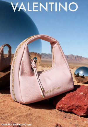 Borsa Valentino rosa tenue con logo dorato e dettaglio catena, posata sulla sabbia del deserto e rocce rosse, con sfere argentate riflettenti e montagne sullo sfondo.