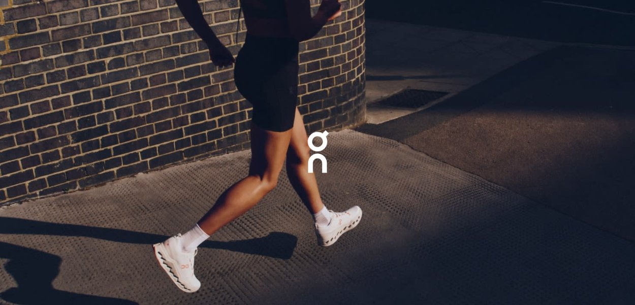 White running shoes with patterned soles, worn with black shorts, while jogging on a textured pavement against a brick wall background.