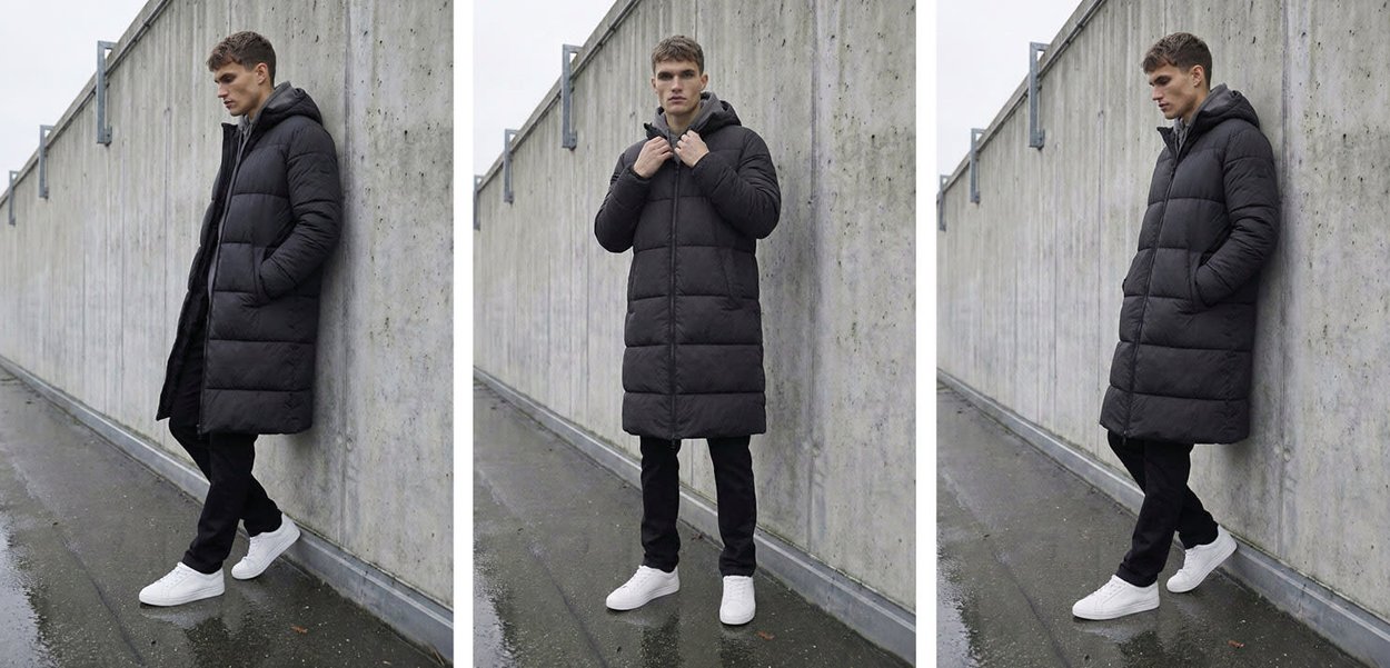 Giovane uomo con cappotto imbottito nero lungo, pantaloni neri e sneakers bianche, in piedi e appoggiato a un muro di cemento all'aperto.