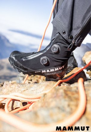 Gros plan sur la chaussure d'alpinisme noire Mammut d'un grimpeur avec des boutons BOA sur un terrain rocheux, des cordes d'escalade orange en arrière-plan.
