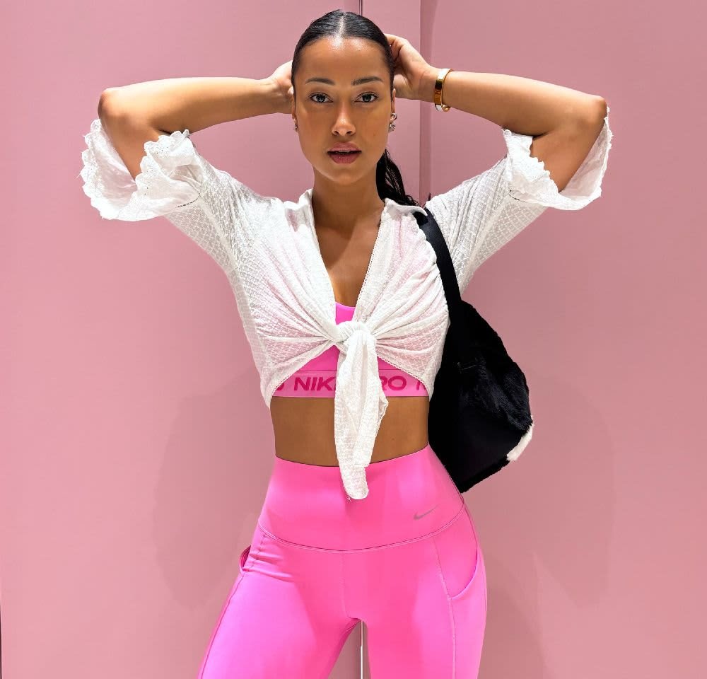 White textured wrap top, pink athletic bra, and high-waisted pink leggings. Black tote bag on shoulder. Background is a solid pink wall.