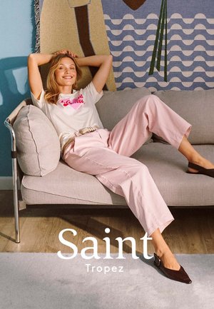 Femme se détendant sur un canapé gris clair, portant un t-shirt blanc à motif, un pantalon rose et des ballerines pointues foncées, avec une tenture murale abstraite colorée en arrière-plan.