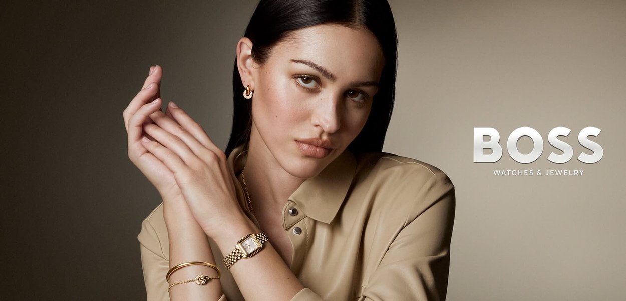 Femme aux cheveux foncés portant une montre en or, un bracelet et des boucles d'oreilles, vêtue d'un haut beige, posant avec les bras croisés devant un fond neutre.