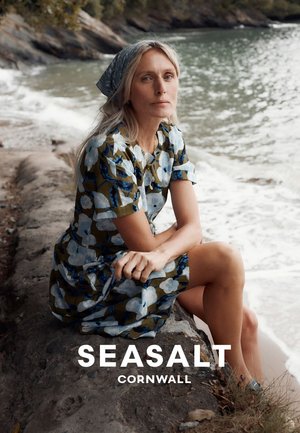 Femme en robe fleurie et foulard assise sur une rive rocheuse, regardant la caméra avec la mer et les vagues en arrière-plan.