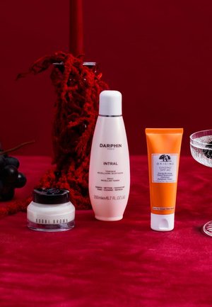 Drei Hautpflegeprodukte vor einem roten Hintergrund: eine klare Flasche mit Flip-Deckel, ein weißes Glas mit einem schwarzen Deckel und eine orangefarbene Tube mit einem weißen Deckel.