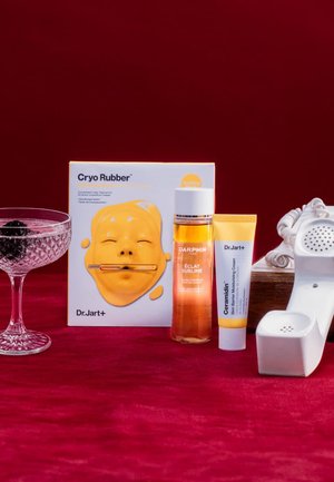 Gelbe Silikon-Gesichtsmaske, Glasbehälter mit Brombeeren, bernsteinfarbenes Öl in einer klaren Flasche, weiße Cremetube, auf strukturiertem roten Hintergrund.