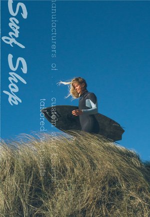 Een persoon in een zwarte wetsuit met lichtblauwe mouwen staat op gras en houdt een donker surfboard vast tegen een heldere blauwe lucht.