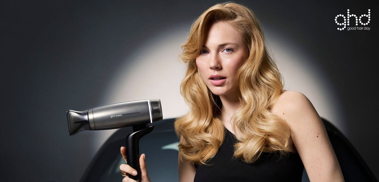 Femme blonde aux cheveux bouclés portant une tenue noire, appuyée sur une voiture noire brillante, tenant un sèche-cheveux argenté, logo ghd visible au-dessus.