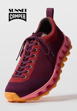 Zapatilla de punto en púrpura y rojo con suela segmentada rosa y puntera naranja, con cordones púrpuras y una lengüeta en el talón.