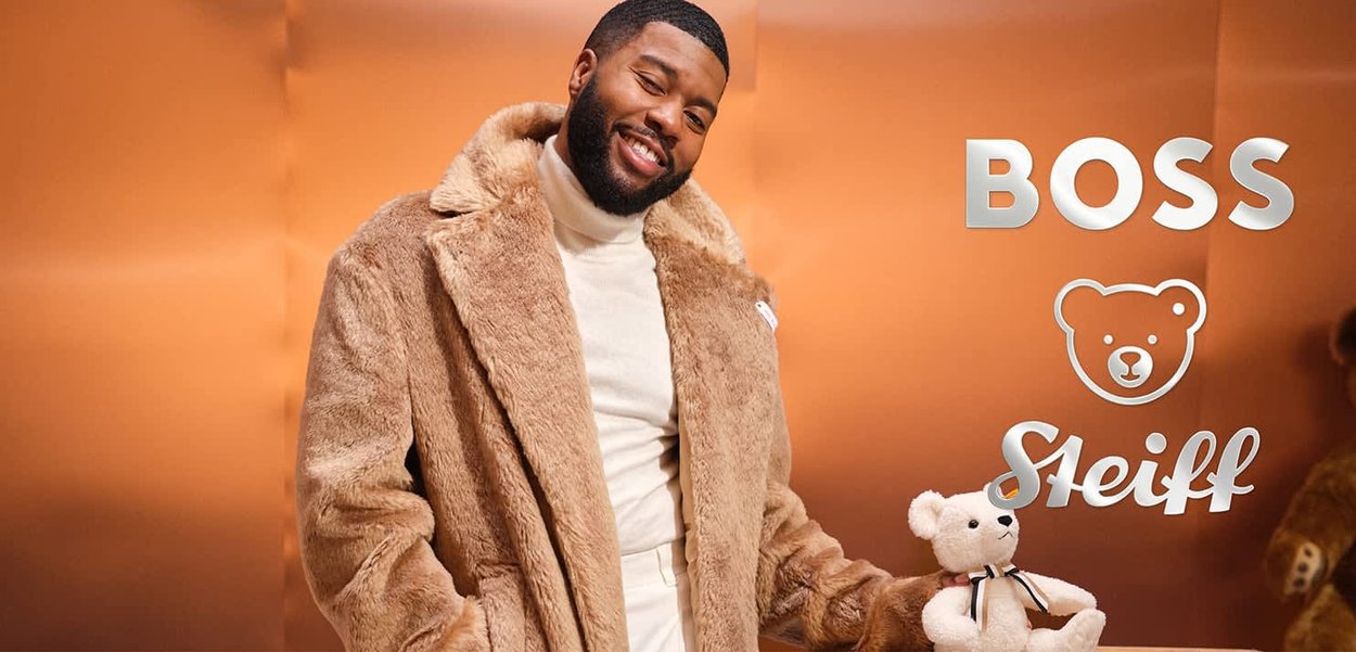 Brauner Kunstfellmantel mit breiten Revers, getragen über einem cremigen Rollkragenpullover. Hält einen kleinen beigen Teddybär mit einem schwarzen Band.