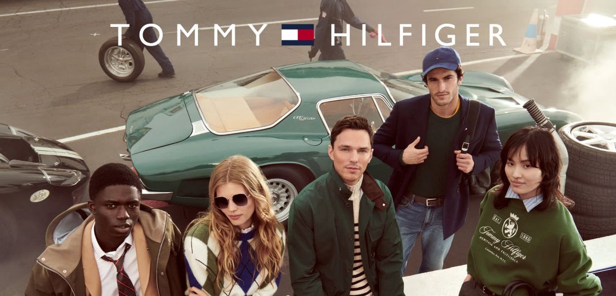 Group of four individuals in casual attire posing in front of a vintage green car. Notable details include sunglasses, a striped sweater, and a logo sweatshirt.