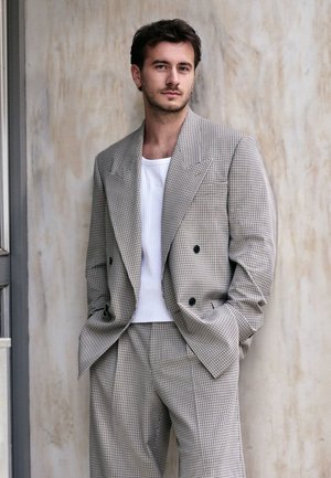 Hombre con cabello corto y oscuro y pendientes, vestido con un traje gris a cuadros y camisa blanca de canalé, de pie con las manos en los bolsillos frente a una pared beige.