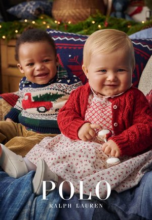 Due bambini siedono vicini l'uno all'altro. Uno indossa un maglione a righe verdi e rosse con un camion, e l'altro un cardigan rosso su un vestito con fantasia.