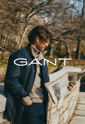 Man in marineblauwe jas en beige sweater leest buiten een krant bij een stenen muur met kale bomen op de achtergrond.