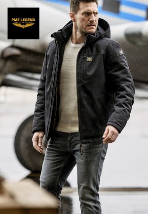 Chaqueta negra con capucha y cierre frontal con cremallera, con un logo sutil en el pecho. Lleva puesta sobre un suéter blanco texturizado y vaqueros azul claro.