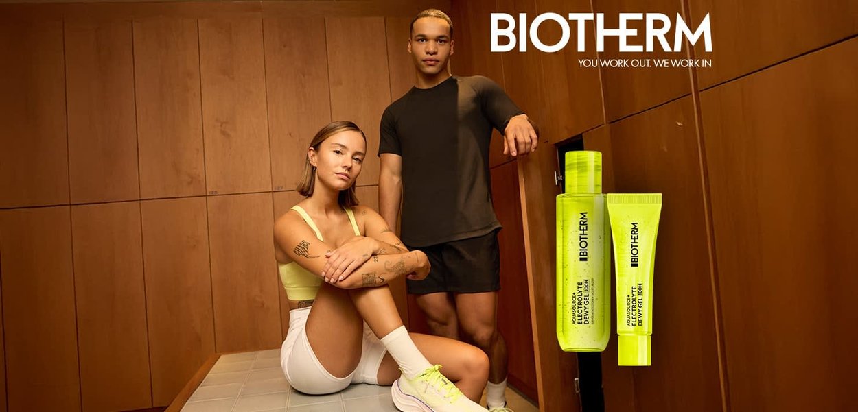 Junger Mann und junge Frau in Sportkleidung posieren in einem hölzernen Umkleideraum neben zwei leuchtend gelben Biotherm Hautpflegeprodukten.