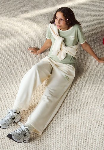 Hellgrünes T-Shirt mit Text, cremefarbene oversized Jogginghose und graue Sportschuhe. Strukturierten Hintergrund. Lässiges Outfit mit geschichteten Kleidungsstücken.