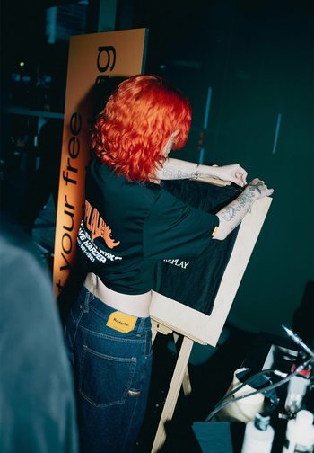 Person mit leuchtend roten Haaren und Tattoos, der ein schwarzes Shirt auf einer Holzstaffelei in einem schwach beleuchteten Innenraum anpasst.