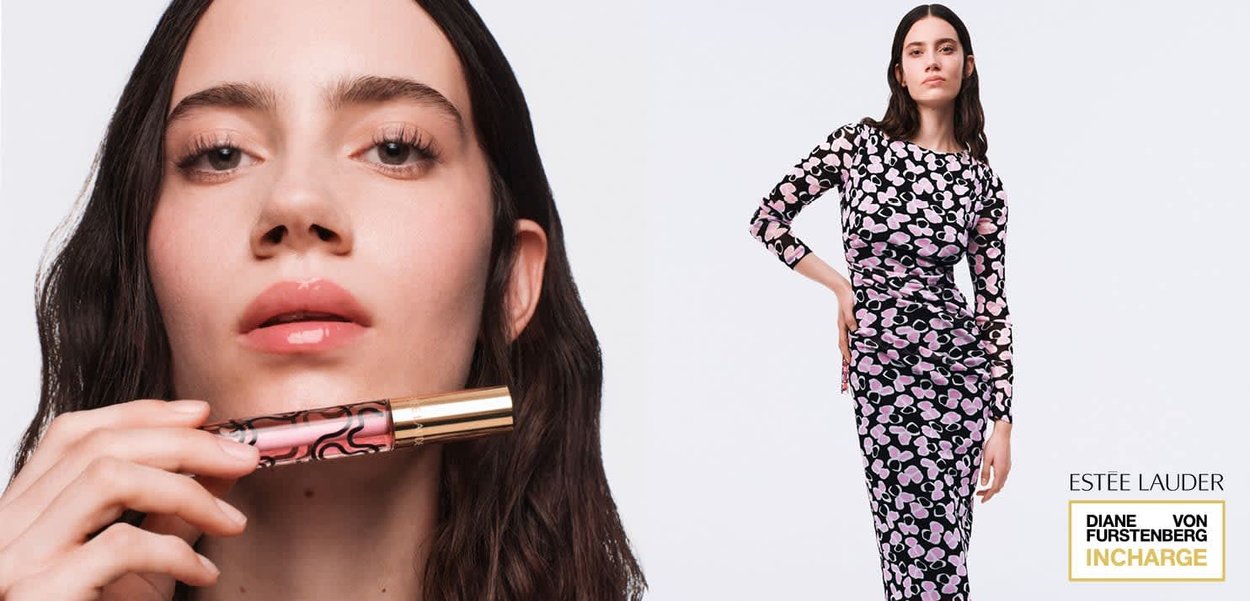 Donna con capelli lunghi e scuri che tiene un lucidalabbra rosa vicino alle labbra, indossa un abito aderente a maniche lunghe nero con motivo rosa, in piedi davanti a uno sfondo bianco.