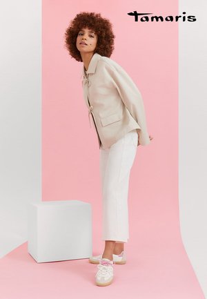 Frau mit lockigem Haar, die eine beige Jacke, weiße Cropped-Hosen und beige Turnschuhe mit pinken Schnürsenkeln trägt und sich vor einem rosa-weißen Hintergrund nach vorne lehnt.