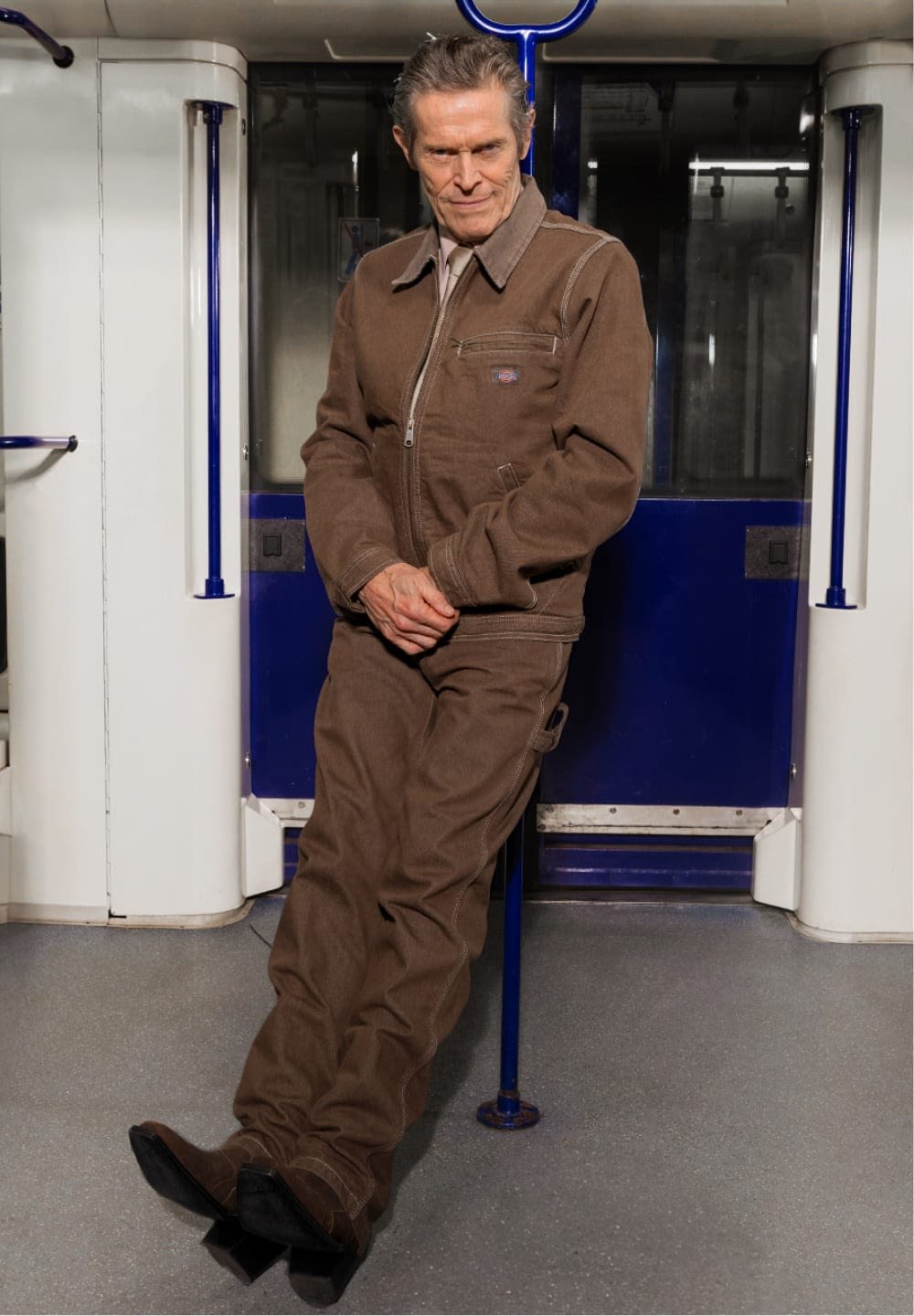 Giacca da lavoro marrone e pantaloni abbinati realizzati in materiale robusto. Presenta una cerniera e tasche, indossati con scarpe nere. Lo sfondo include un ambiente urbano.