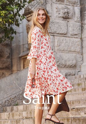 Jeune femme en robe blanche à fleurs rouges tenant des lunettes de soleil, montant des escaliers en pierre en plein air près d'un vieux mur de pierre.