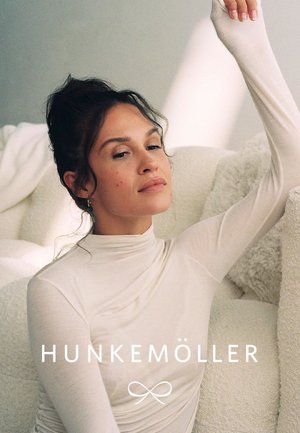 Een model draagt een nauwsluitend, lang mouw, crèmekleurig top. De achtergrond heeft een zachte, gestructureerde, lichtgekleurde stof. Logo onderaan.