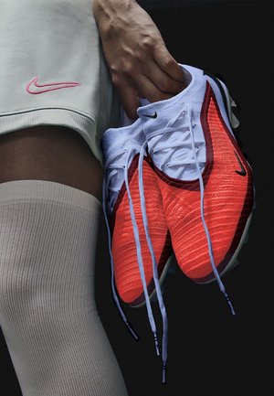 Scarpe da ginnastica Nike, con una colorazione rosso e bianco, tomaia testurizzata, dettagli neri e lacci azzurri chiari, sorrette da una mano.