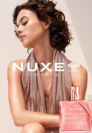 Kobieta z krótkimi kręconymi włosami, ubrana w lśniącą top na halter, siedzi obok różowej butelki wielofunkcyjnego suchego oleju NUXE.