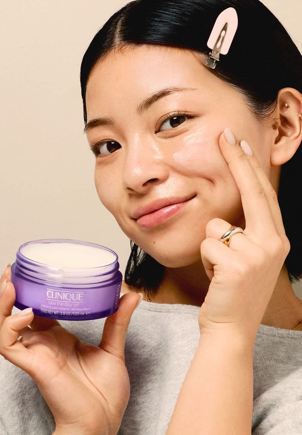 Vrouw met zwart haar brengt met vinger Clinique Take The Day Off reinigingsbalsem aan op haar wang, terwijl ze een open paarse pot in haar hand houdt.