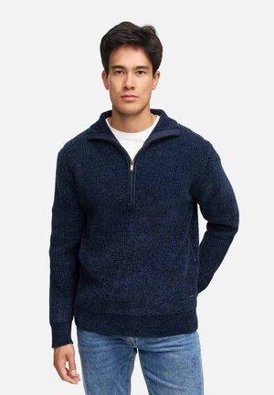 Giovane uomo che indossa un maglione a mezza cerniera blu navy a coste sopra una camicia bianca e jeans azzurri, in piedi contro uno sfondo semplice.