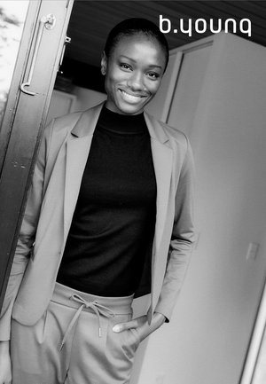 Hellgrauer Blazer über einem schwarzen Rollkragenpullover, kombiniert mit grauen Hosen. Die Person steht an einer offenen Tür und lächelt sanft.